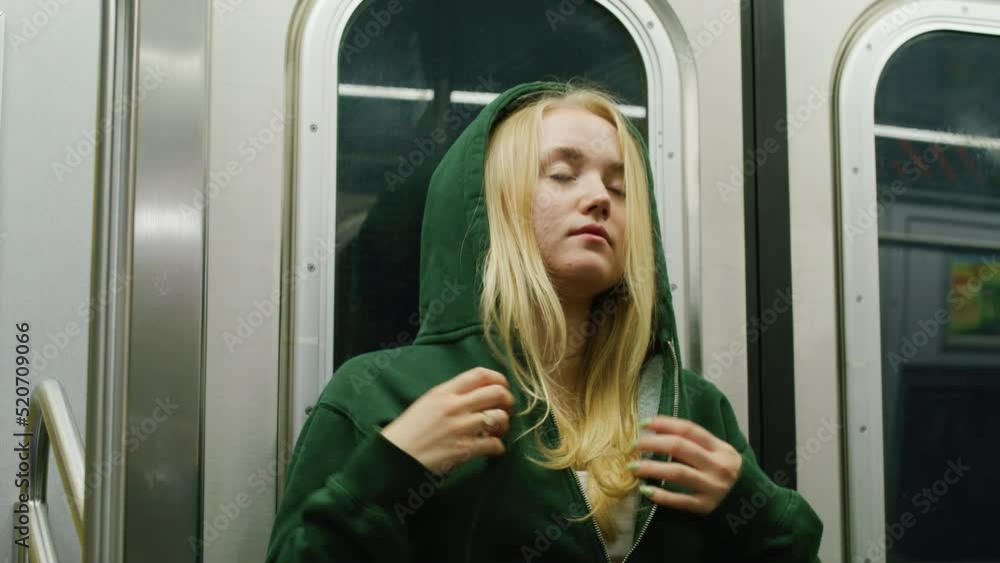 Portrait of a young teen blonde woman passenger in headphones, standing in underground train public transport subway. Girl putting on hood of Emerald green hoodie. Listening to music. 4k Portrait of a young teen blonde woman passenger in headphones, standing in underground train public transport subway. Girl putting on hood of Emerald green hoodie. Listening to music. 4k
