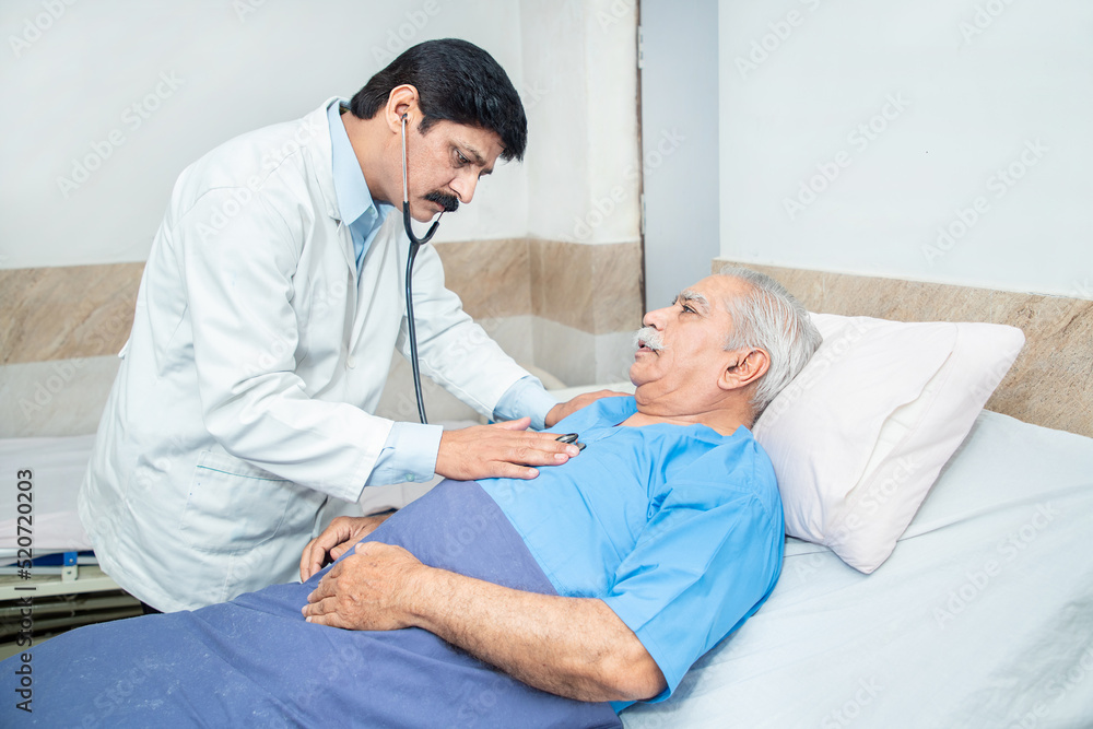 Indian doctor wearing stethoscope check heart beat of olde senior male patient lying on bed at clinic or hospital. Elderly people health care concept. Indian doctor wearing stethoscope check heart beat of olde senior male patient lying on bed at clinic or hospital. Elderly people health care concept.