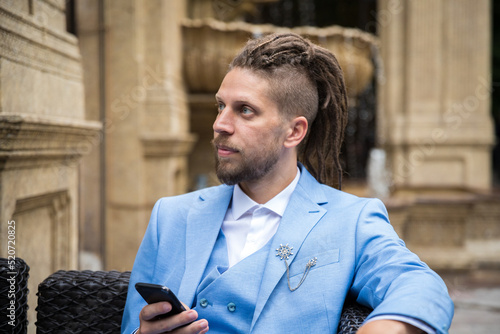 Canvas-taulu Young successful hipster caucasian man with dreadlocks in a blue suit with a smartphone in his hands sits in a street restaurant looks away