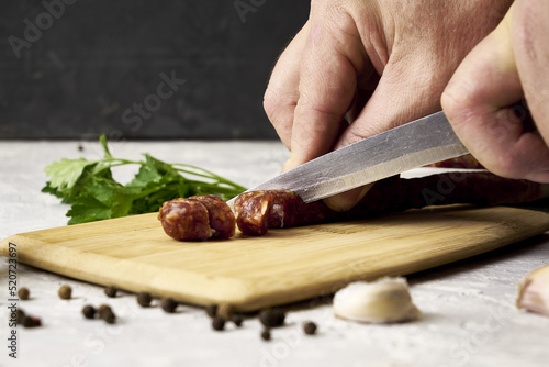 a man cuts spicy smoked sausages on a cutting board next to red pepper garlic spices and herbs on the table