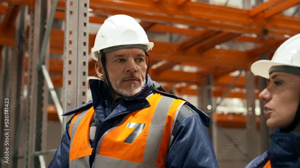 Experienced engineers discuss working issues walking near empty metal racks in warehouse. Man and woman in white hardhats talk closeup slow motion Experienced engineers discuss working issues walking near empty metal racks in warehouse. Man and woman in white hardhats talk closeup slow motion