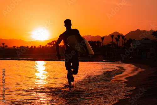 Silhouette man surfer in sunset light running down beach with surfboard near waters edge enjoying summer time practice hobby during getaway vacations, guy recreating at tropical island with waves