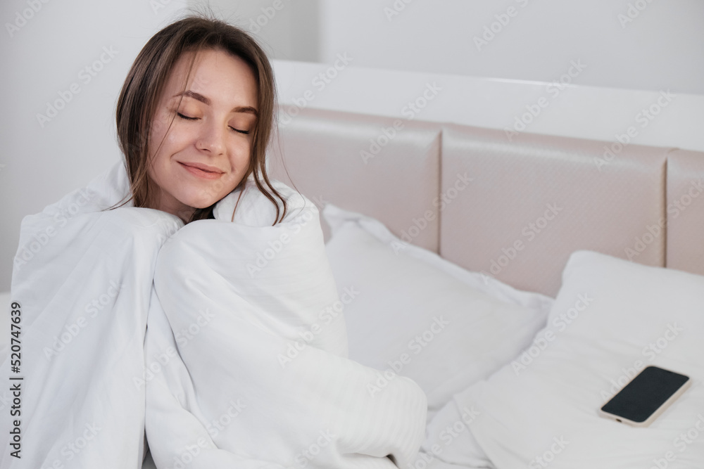 Pretty young smiling Caucasian woman waking up in the morning and stretching in her bed and looking content. Covered in white linen. Pretty young smiling Caucasian woman waking up in the morning and stretching in her bed and looking content. Covered in white linen.
