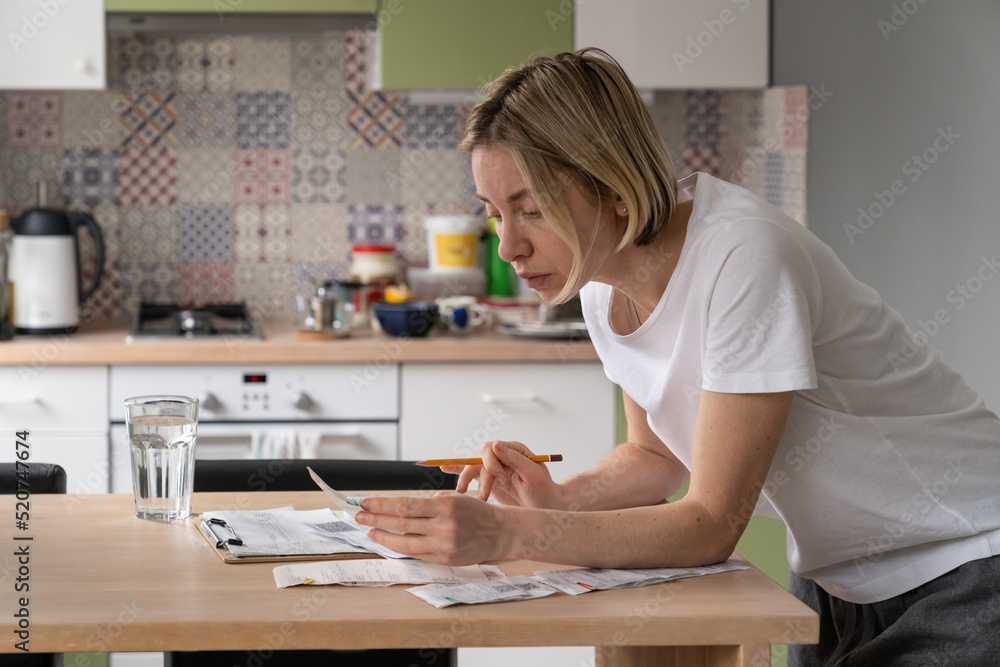 Mature woman looks at communal payments bills placed on table. Thoughtful single female of middle age looks unsatisfied with utility bill sums. Blonde lady examines papers in kitchen closeup Mature woman looks at communal payments bills placed on table. Thoughtful single female of middle age looks unsatisfied with utility bill sums. Blonde lady examines papers in kitchen closeup