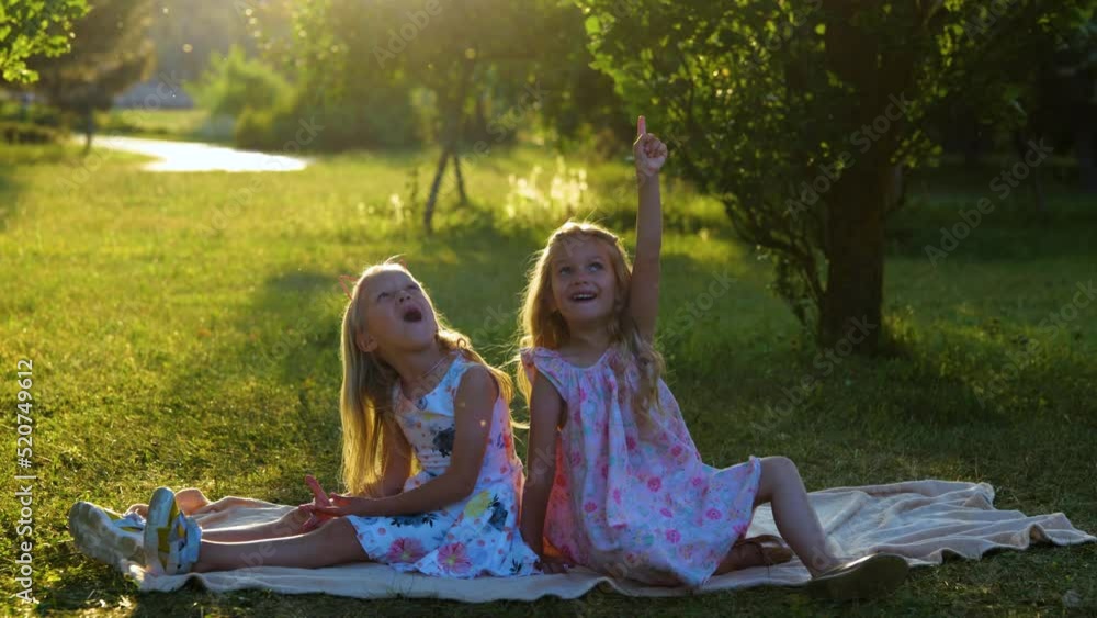 Happy little girls sitting on a blanket in park and looking up with amazed faces. Caucasian blonde carefree children outside having fun playing on nature. Happy childhood concept Happy little girls sitting on a blanket in park and looking up with amazed faces. Caucasian blonde carefree children outside having fun playing on nature. Happy childhood concept