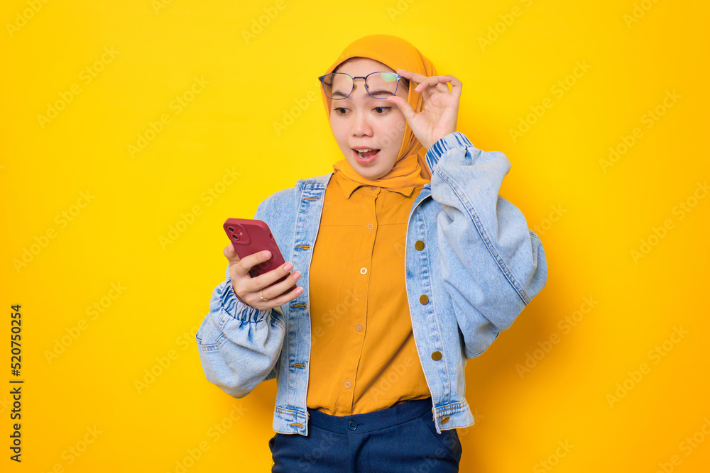 Shocked young Asian woman in jeans jacket looking at mobile phone screen, taking off glasses with opening mouth, reacting to great sale offers isolated over yellow background