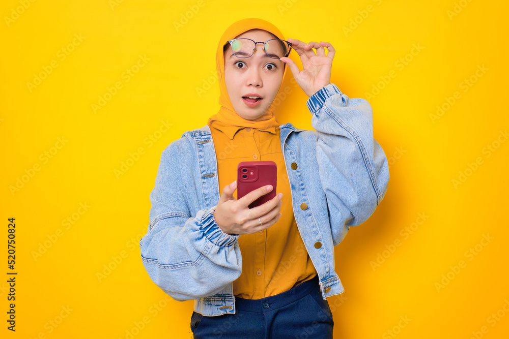 Shocked young Asian woman in jeans jacket holding mobile phone, taking off glasses with opening mouth, reacting to great sale offers isolated over yellow background Shocked young Asian woman in jeans jacket holding mobile phone, taking off glasses with opening mouth, reacting to great sale offers isolated over yellow background