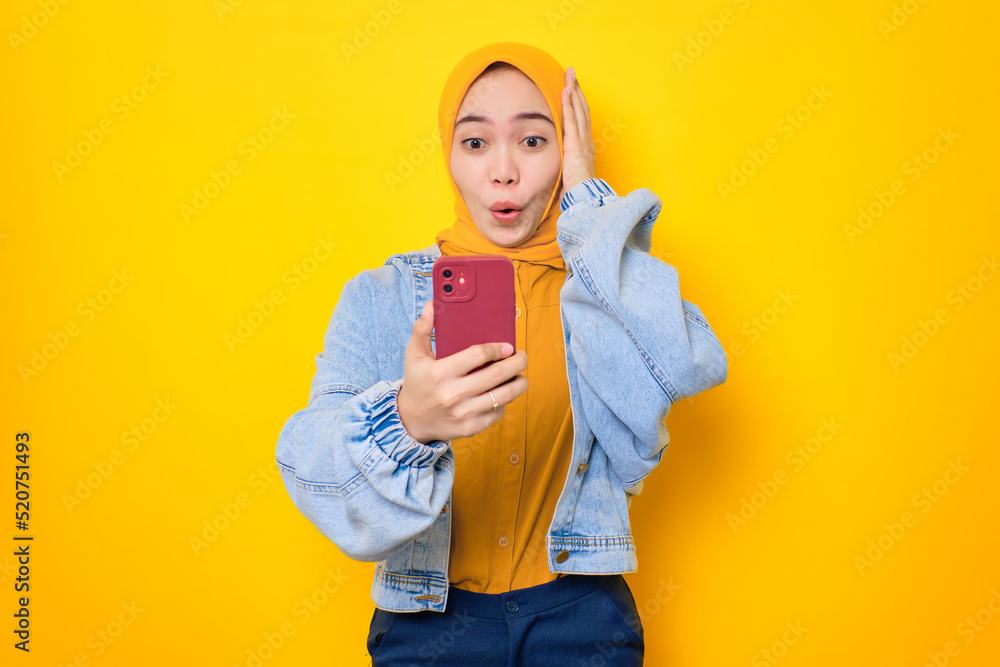 Shocked young Asian woman in jeans jacket looking at mobile phone screen with opening mouth, reacting to great news isolated over yellow background Shocked young Asian woman in jeans jacket looking at mobile phone screen with opening mouth, reacting to great news isolated over yellow background