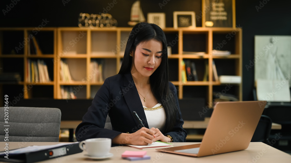 Asian female manager working with laptop and writing her business plan on notepad Asian female manager working with laptop and writing her business plan on notepad