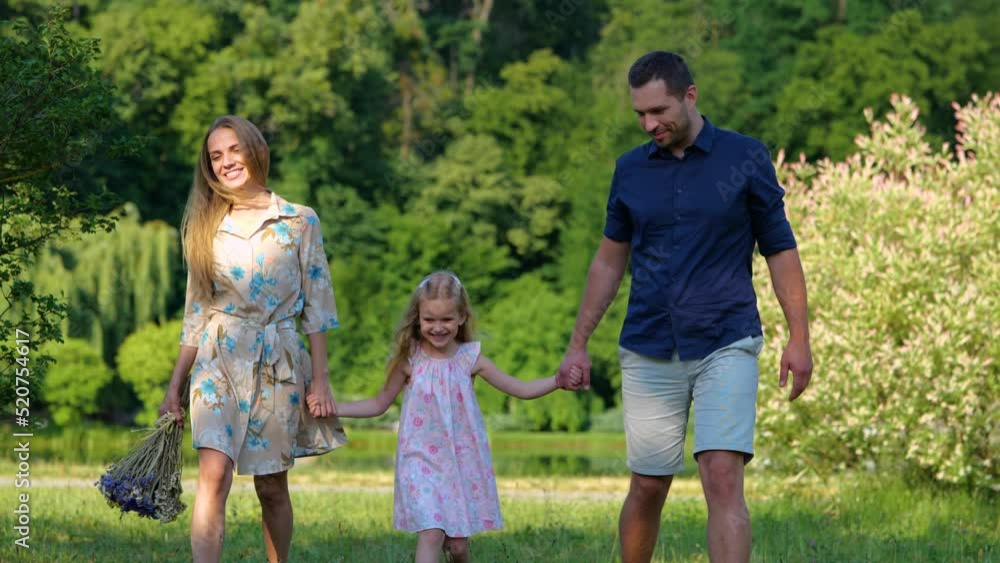 Positive family walking outdoor with child. Happy beautiful mother with flowers with daughter and husband on nature. Parents holding hands with little girl enjoying walk and smiling Positive family walking outdoor with child. Happy beautiful mother with flowers with daughter and husband on nature. Parents holding hands with little girl enjoying walk and smiling
