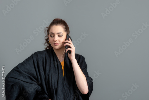 curly-haired emotional young woman in a black cloak talk on mobile phone, isolated on gray