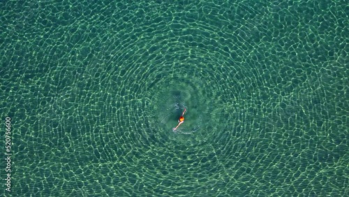 Aerial view a woman in an open, clear, transparent sea is spinning, creating round waves around her, a lot of glare and reflexes from the water, shooting from a drone
