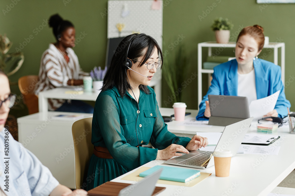 Asian young operator typing on laptop while talking with customer using headphones with other managers around her Asian young operator typing on laptop while talking with customer using headphones with other managers around her