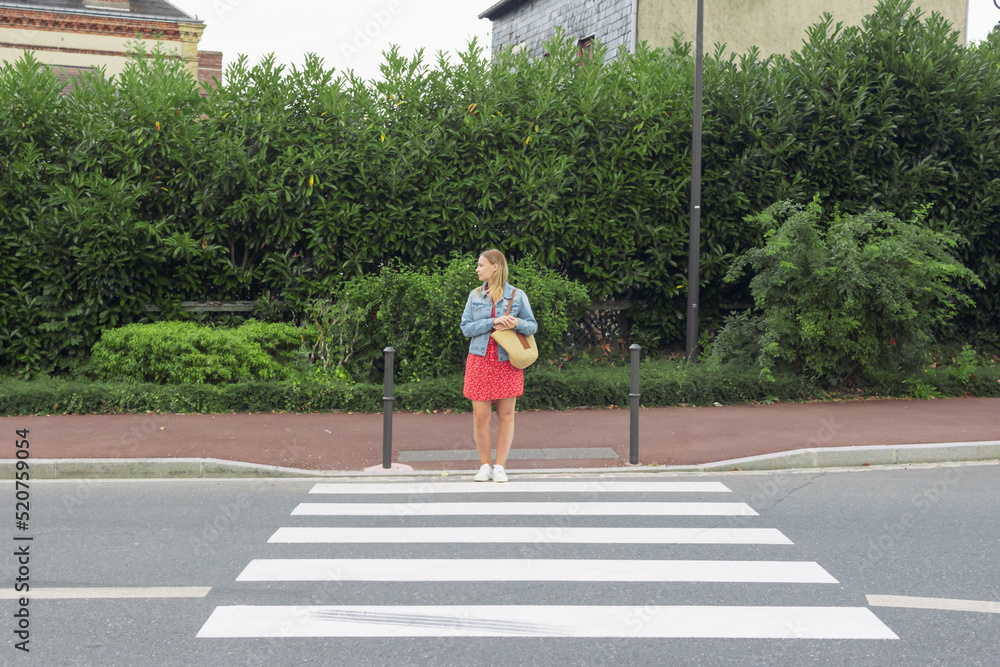 A girl in a red dress and a denim jacket with a bag on her shoulder stands at a pedestrian crossing and looks to cross the road