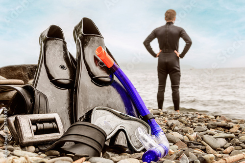 Flippers mask and cargo for scuba diving lie on the rocky seashore. In the background, a male diver stands with his back to the camera and looks towards the ocean. photo