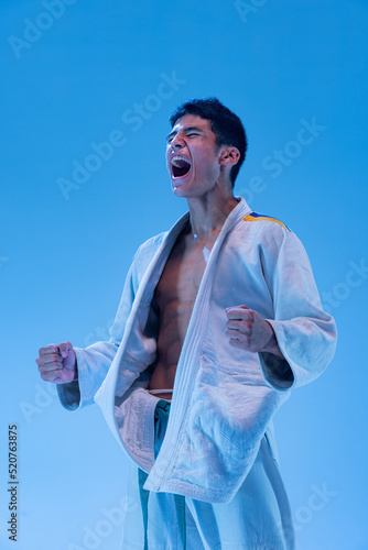 Slika na platnu Portrait of young muscular man in white kimono screaming, posing isolated over blue studio background in neon light