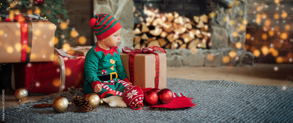 Little child under Christmas tree. baby girl in Santa Claus hat with gifts under Christmas tree with many gift boxes presents. Happy Holidays, New year. Cozy warm winter evening at home. Xmas time