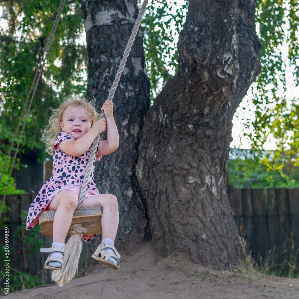 A baby girl in a pink dress swings on a bungee swing made of a wooden board with a rope attached to a tree branch in summer or spring