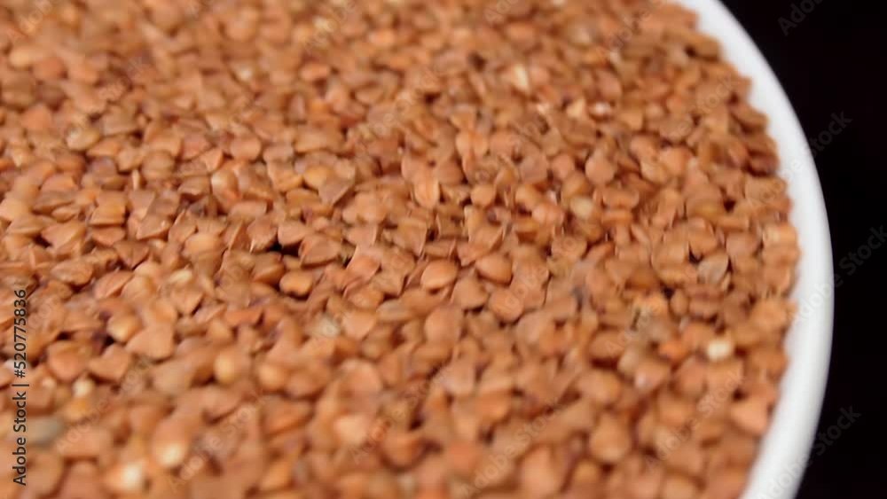Dry Uncooked Brown Buckwheat Groats on White Plate Rotating against Black Background. Raw Large Buckwheat Grains. Russian, Ukrainian and Belarusian Culture Dry Uncooked Brown Buckwheat Groats on White Plate Rotating against Black Background. Raw Large Buckwheat Grains. Russian, Ukrainian and Belarusian Culture