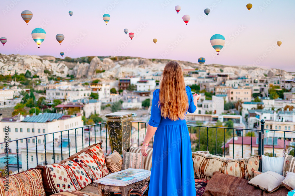 young woman in blue dress stands on terrace of hotel in Goreme Cappadocia and looks at hot air balloons rising into sky, concept  of must see travel destination, bucket list trip