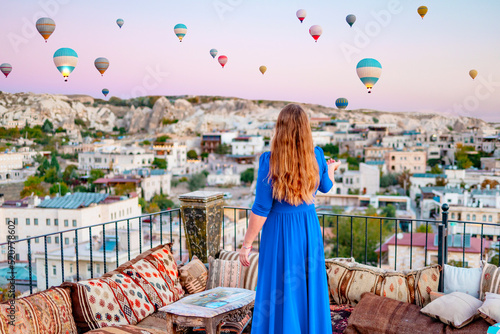 young woman in blue dress stands on terrace of hotel in Goreme Cappadocia and looks at hot air balloons rising into sky, concept  of must see travel destination, bucket list trip photo