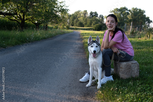 A young woman sits with her husky dog on the side of a village road and waits for a car for hitch-hiking © Diana