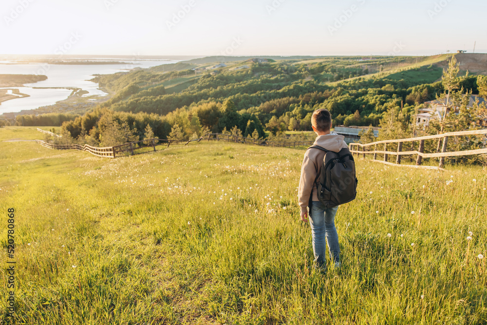 Tourist with backpack standing on top of hill in grass field and enjoying beautiful landscape view. Rear view of teenage boy hiker resting in nature. Active lifestyle. Concept of local travel