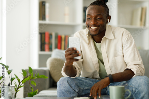  African American man in headphones using a smartphone, browsing the Internet while sitting on the sofa at home, writing on a mobile phone or using a new application indoors