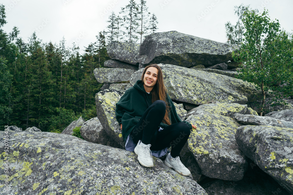 Attractive happy female tourist sitting on a rock in the mountains and posing for the camera with a smile on her face.