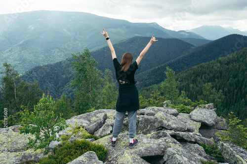 A female tourist in casual clothes with raised hands stands on a rock and looks at the beautiful landscape of the Carpathians