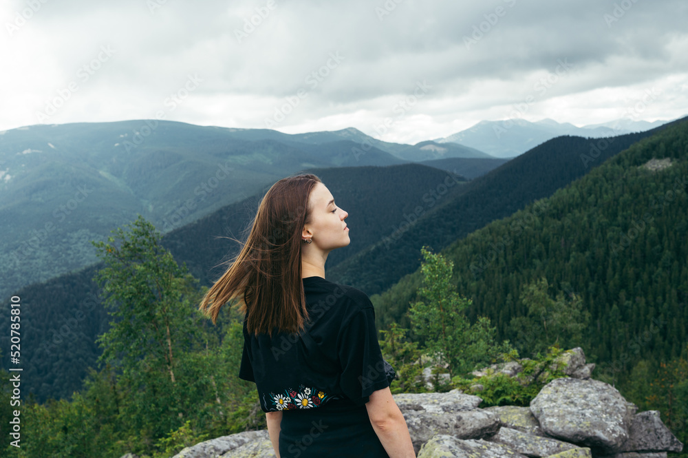 Portrait of a stylish lady tourist in casual clothes on top of a mountain standing on a rock and posing for the camera against the background of mountain views.