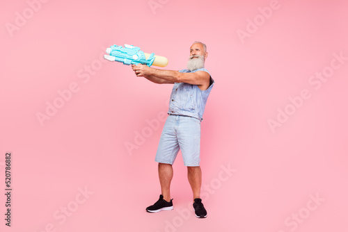 Full length photo of cool handsome retired guy wear jeans waistcoat shooting water weapon isolated pink color background