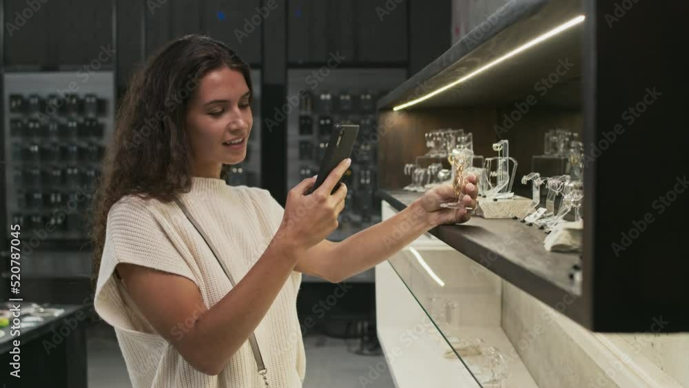 Waist-up of young brown-haired woman taking picture with smartphone of earrings in contemporary jewelry store
