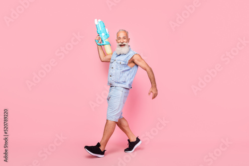 Full length photo of funny charming man pensioner dressed denim vest walking rising water gun isolated pink color background