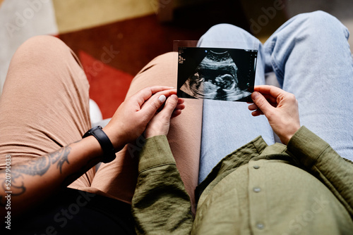 Close up of tattooed gay couple with pregnant young woman looking at ultrasound image of baby, same sex family photo