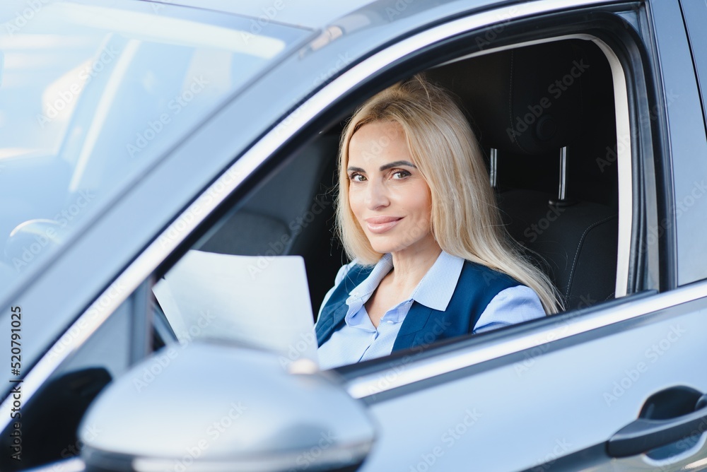 Portrait of a beautiful mature business woman driving a car, sitting behind steering wheel, looking at camera and smiling