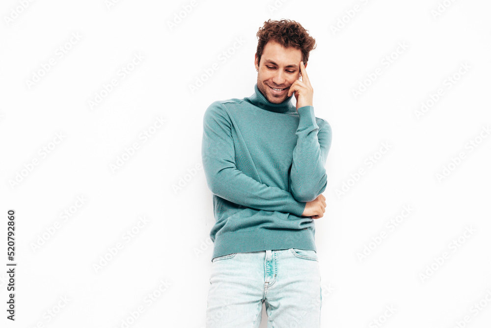 Fototapeta premium Portrait of handsome confident model. Sexy stylish man dressed in blue sweater and jeans. Fashion hipster male with curly hairstyle posing near white wall in studio. Isolated