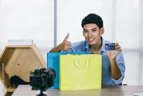 A young Asian influence male vlogger holding a credit card with shopping bags on his desk on live streaming, youtuber concept.