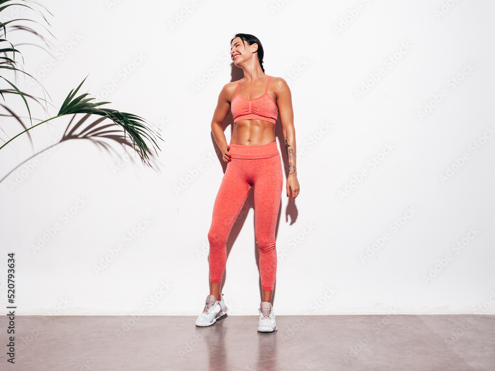 Tanned fitness woman in pink sports clothing. Sexy young smiling beautiful model with perfect body.Female isolated on white background in studio. Stretching out before training. Near wall. Full length