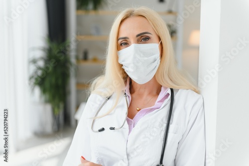 Middle age blonde woman wearing doctor uniform and medical mask standing at clinic