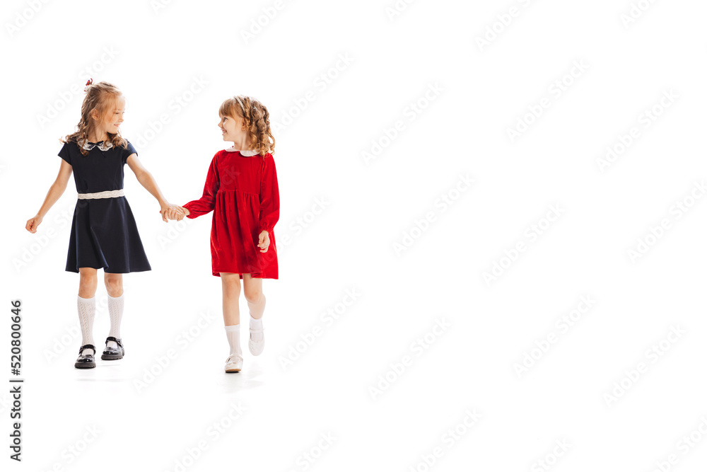 Portrait of two little girls, children in beautiful dresses holding hands, cheerfully walking isolated over white studio background Portrait of two little girls, children in beautiful dresses holding hands, cheerfully walking isolated over white studio background