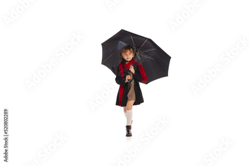 Portrait of beautiful little girl, child in stylish retro outfit posing, walking under umbrella isolated over white studio background