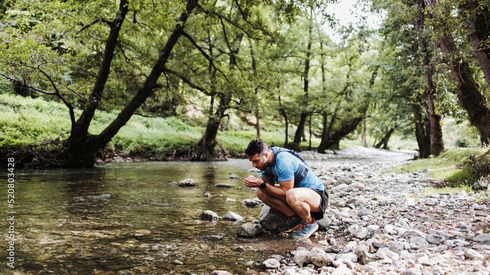 Young adult sporty man fitness and triathlon training in nature. He is drinking water from the fresh mountain river and preparing himself for a race or training. People and extreme sports concept. Young adult sporty man fitness and triathlon training in nature. He is drinking water from the fresh mountain river and preparing himself for a race or training. People and extreme sports concept.