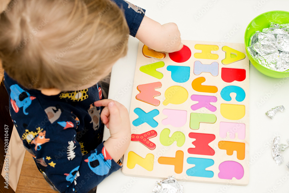 Two year boy playing with wooden alphabet letters board. Letters wrapped in foil. Intellectual game, preschool implement for early education. Verbal and memory training exercise. Two year boy playing with wooden alphabet letters board. Letters wrapped in foil. Intellectual game, preschool implement for early education. Verbal and memory training exercise.
