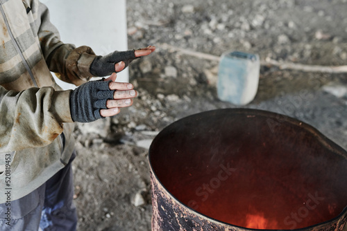 Quadro su tela Close-up of poor man in torn gloves and dirty clothes warming up by fire standin