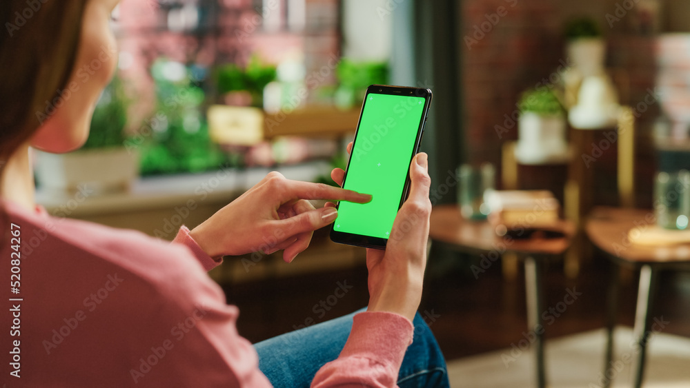 Feminine Hand Scrolling Feed on Smartphone with Green Screen Mock Up Display. Female is Resting at Home and Checking Social Media on Mobile Device. Close Up Over the Shoulder Photo.