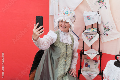 Smiling senior woman in stylized ancient Russian headdress - kokoshnik taking selfie on cell phone. photo