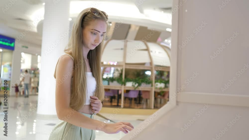 A beautiful young girl using a touch terminal in a shopping mall.The girl uses a touch terminal to search for the right store.The concept of people and technologies. Airport, online booking. Router A beautiful young girl using a touch terminal in a shopping mall.The girl uses a touch terminal to search for the right store.The concept of people and technologies. Airport, online booking. Router