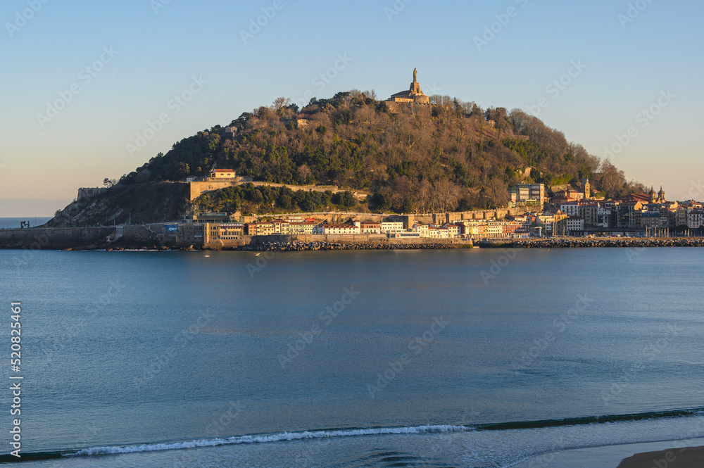 Naklejka premium view of monte urgull, the old port of San Sebastián, the old city, at sunrise on a lonely winter day. Bay with the sea completely calm, in the foreground small waves breaking on the sand