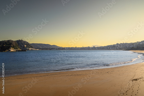 View of the sunrise in San Sebastian from Ondarreta beach on a winter day at low tide. The beach in the foreground, the calm sea, some waves, and in the background Mount Urgull and the city at its fee